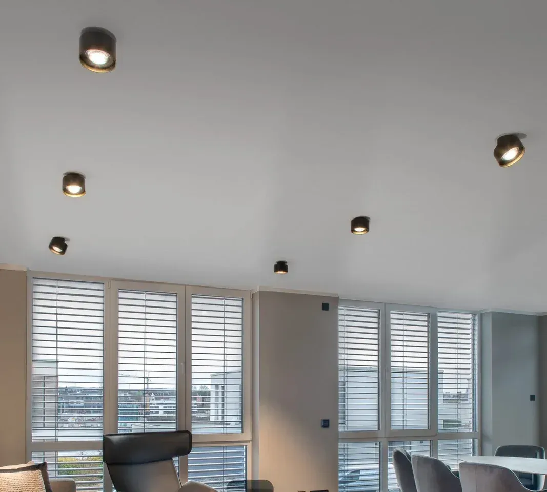 Wohnraum mit Fly Glas Aufbauspots in Rauchglas an der hellen Decke vor Fenstern mit Jalousien Wohnraum mit Fly Glas Aufbauspots in Rauchglas an der hellen Decke vor Fenstern mit Jalousien